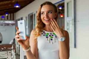 attraktiv Frau im Weiß Kleid im Sommer- Cafe im Sonnenbrille mit Telefon nehmen Foto