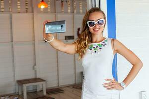 attraktiv Frau im Weiß Kleid im Sommer- Cafe im Sonnenbrille mit Telefon nehmen Foto
