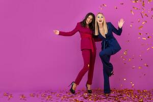 zwei attraktiv Frauen feiern Neu Jahr auf violett Hintergrund im stilvoll bunt Abend Anzüge von lila und Blau Farbe, freunde haben Spaß zusammen, Mode Trend, golden Konfetti Party Stimmung foto