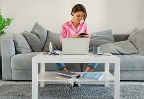 Frau im Rosa Hemd Sitzung entspannt auf Sofa beim Zuhause beim Tabelle Arbeiten online auf Laptop foto