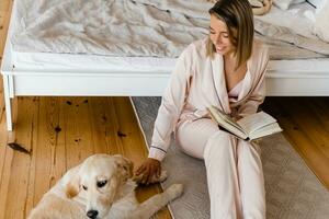 ziemlich lächelnd Frau beim Zuhause im Morgen im Pyjama lesen Buch spielen mit Hund foto