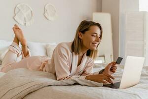 ziemlich lächelnd Frau Einkaufen online auf Laptop beim Zuhause auf Bett im Morgen im Pyjama foto