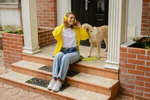 Frau im Gelb Sweatshirt Gehen beim ihr Haus mit ein Hund Hören zu Musik- im Kopfhörer foto