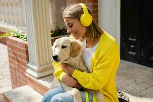 Frau im Gelb Sweatshirt Gehen beim ihr Haus mit ein Hund Hören zu Musik- im Kopfhörer foto