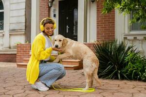 Frau im Gelb Sweatshirt Gehen beim ihr Haus mit ein Hund Hören zu Musik- im Kopfhörer foto