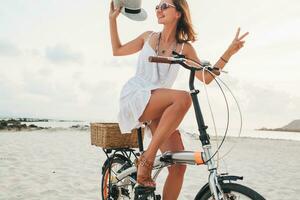 jung attraktiv lächelnd Frau im Weiß Kleid Reiten auf tropisch Strand auf Fahrrad foto