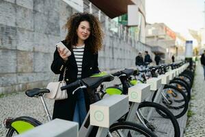 ziemlich lockig Frau mieten ein Fahrrad im Straße mit ein App foto