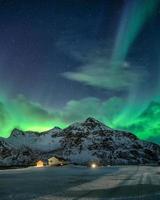 Aurora Borealis mit Sternenhimmel über schneebedeckten Bergen und nordischem Dorf nachts in Flakstad, Lofoten-Inseln