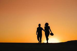 Silhouetten eines glücklichen jungen Paares auf einem Hintergrund des orangefarbenen Sonnenuntergangs in der Sandwüste foto