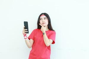 Porträt von schön asiatisch Frau tragen rot Outfit feiern Indonesien Unabhängigkeit Tag halten Handy, Mobiltelefon Telefon mit Denken Ausdruck foto