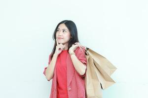 Porträt von schön asiatisch Frau tragen rot Outfit gestikulieren Tragen viele von Einkaufen Taschen mit Denken Ausdruck foto