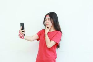 Porträt von schön asiatisch Frau tragen rot Outfit feiern Indonesien Unabhängigkeit Tag halten Handy, Mobiltelefon Telefon mit Denken Ausdruck foto