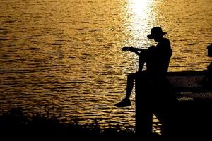 Silhouette des Gitarristen, der eine Gitarre auf dem Fluss unter dem Sonnenuntergang spielt foto
