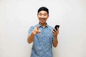 positiv asiatisch Mann Blau Hemd halten Handy, Mobiltelefon Telefon Lächeln und Punkt Finger beim Sie isoliert foto