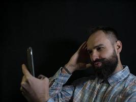 stylischer Mann mit Bart und Schnurrbart posiert und macht ein Selfie am Telefon auf schwarzem Hintergrund