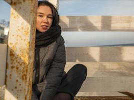 Attraktives junges Mädchen mit zerzausten Haaren sitzt in einem Mantel auf einem Holzdesign am Strand foto