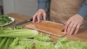 Nahaufnahme von Mannhänden, die Gemüse auf Holzbrett für Salat auf dem Tisch mit gesundem Essen in der Küche schneiden.