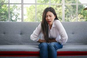 asiatisch lange behaart schön Frau hat ein Magenschmerzen sitzen auf das Sofa im das Leben Zimmer beim heim. Krankenhaus Konzept Frau Krankheit Warnung Zeichen von verschiedene Krankheiten oder Bauch Schmerzen Vor Menstruation. foto