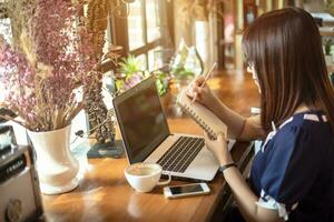 asiatisch Geschäft weiblich Arbeiten mit machen ein Hinweis mit ein Notizbuch und Laptop im Kaffee Geschäft mögen das Hintergrund. foto