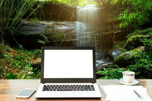 Attrappe, Lehrmodell, Simulation Bild von Laptop mit leer Weiß Bildschirm, intelligent Telefon und dokumentieren auf hölzern Tabelle beim Wasserfall im tropisch Regenwald Landschaft, Freizeit und Reise Konzept. foto