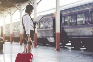 asiatisch Frau schwanger Reise aussehen beim das Karte mit rot Koffer beim Eisenbahn Bahnhof Reisen, Urlaub Reise Konzept. foto