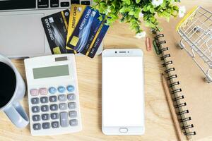 Anerkennung Karte im Einkaufen Wagen mit Laptop, Notizbuch, Blume Topf Baum, Smartphone und Taschenrechner auf hölzern Hintergrund, online Bankwesen Konzept, oben Aussicht Büro Tisch. foto