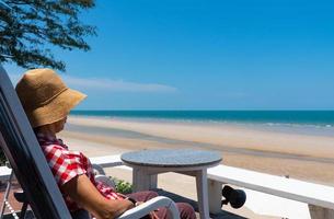 Senior Frau sitzt auf dem Balkon des Hotels mit Blick auf das blaue Meer und den weißen Strand an einem sonnigen Tag der Sommerferien? foto