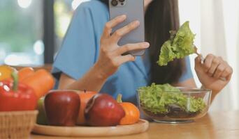 ein jung Frau mit ein schön Gesicht im ein Blau Hemd mit lange Haar Essen Obst Sitzung Innerhalb das Küche beim Zuhause mit ein Laptop und Notizbuch zum Entspannung, Konzept Urlaub. foto