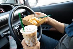 asiatische dame, die eiskaffee und brotbäckerei im auto hält, ist gefährlich und riskiert einen unfall.