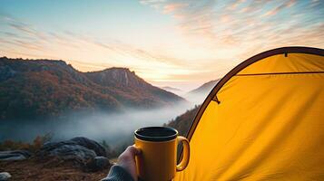 Camping halten ein Tasse im ein öffnen Gelb Zelt mit ein neblig Berg zündete durch natürlich Licht. foto