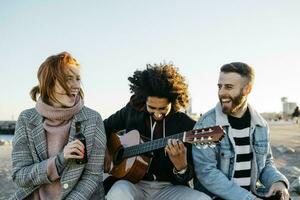 drei glücklich freunde mit Gitarre Sitzung draußen beim Sonnenuntergang foto