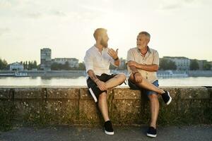 Vater und Erwachsene Sohn Sitzung auf ein Mauer beim das Flussufer reden foto