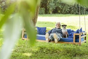 Senior Mann entspannend auf ein schwingen Bett im seine Garten mit seine Hund foto