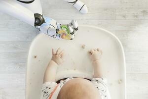 Roboter Hand geben Schnuller zu Baby Junge Sitzung im hoch Stuhl spielen mit Brot Krümel, oben Aussicht foto