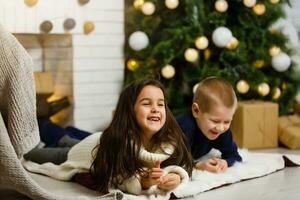 Familie auf Weihnachten Vorabend beim Kamin. Kinder unter Weihnachten Baum mit Geschenk Boxen. dekoriert Leben Zimmer mit traditionell Feuer Ort. gemütlich warm Winter Abend beim heim. foto