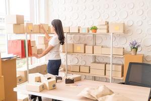 asiatische Geschäftsinhaberin, die zu Hause mit Verpackungsbox am Arbeitsplatz arbeitet - Online-Shopping-KMU-Unternehmer oder freiberufliches Arbeitskonzept