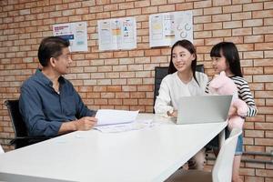 Eine weibliche Managermutter, die mit einem asiatischen männlichen Angestellten am Schreibtisch in einem orangefarbenen Geschäftsbüro mit Ziegelwänden arbeitet und spricht, während ihre Tochter hereinkommt und lächelt.