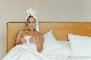 Frau Lügen im Bett halten Kaffee Becher mit ein Handtuch auf ihr Kopf. foto