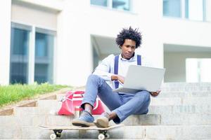 schwarzer Geschäftsmann mit Afrohaar und Skateboard mit seinem Laptop his foto