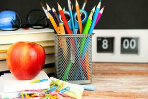 Apfel und Schule liefert auf ein hölzern Schreibtisch. im das Hintergrund, ein Schule Tafel und ein Uhr sind im verwischen. zurück zu Schule. selektiv Fokus. foto