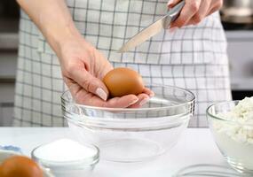 Frau im ein Schürze ist knacken ein Ei in ein Schüssel zum Backen Dessert im das Küche beim heim. Nahansicht. selektiv Fokus. foto