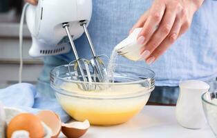ein Frau schlägt Eier mit ein Rührgerät und fügt hinzu Zucker während Stehen im das Küche beim heim. Kochen und Backen Nachspeisen beim heim. selektiv Fokus. foto