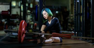 asiatisch Frau ist gegenüber Übertraining Syndrom nach Gewicht Ausbildung trainieren Innerhalb Fitnessstudio mit dunkel Hintergrund zum ausüben und Fitness Konzept foto