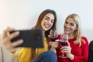 Lachen Frau freunde umarmen jeder andere auf Sofa während nehmen Selfie Foto auf Clever Telefon. liebenswert blond und Brünette Mädchen ausdrücken positiv Emotionen zu Kamera während Trinken Wein