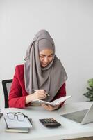 Porträt von schön Muslim Geschäftsfrau lächelnd beim das Kamera, Sitzung beim ihr Schreibtisch. foto