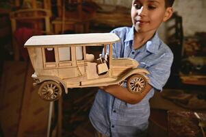 ein Schüler halten ein hölzern Modell- von handgemacht Wagen. Zimmerei, Hobby, Holz Carving Industrie. Fokus auf hölzern Auto foto