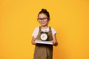 lächelnd wenig Mädchen im Brillen und stilvoll beiläufig Kleid, Tragen ein Buch und Alarm Uhr, isoliert Orange Hintergrund foto