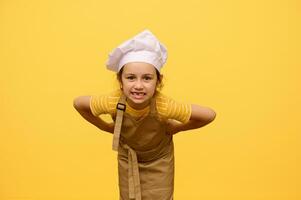 boshaft wenig Mädchen im Koch Hut und Schürze, Grimassen, Herstellung Gesichter, posieren mit Hände auf Taille, isoliert auf Gelb foto