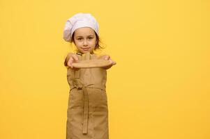 wenig Mädchen im Koch Hut und Schürze, lächelt und hält aus beim Kamera ein hölzern Planke, isoliert Über Gelb Studio Hintergrund foto