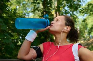 Nahansicht von Sportlerin Trinken Wasser von Flasche, erfrischend und feuchtigkeitsspendend Ihre Körper nach schwer draussen Sport trainieren foto